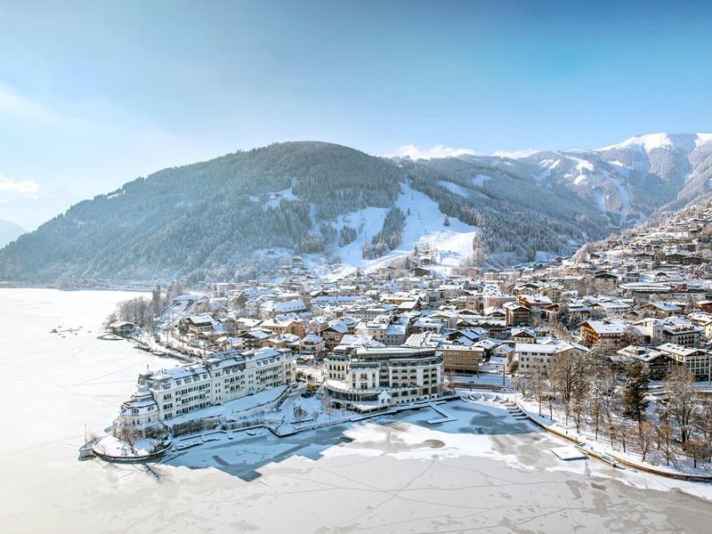 Zasněžená horská vesnice s zamrzlým jezerem a horami v pozadí pod jasnou oblohou