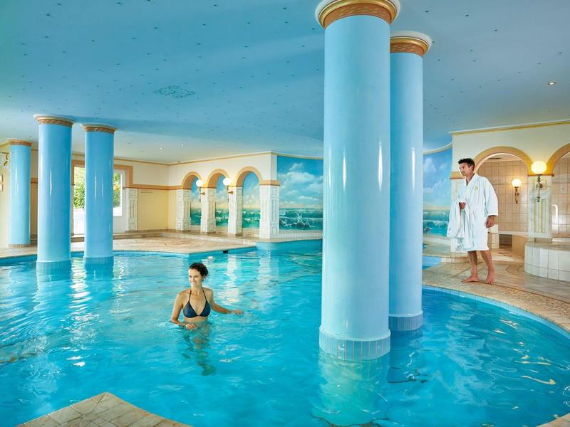 Vnitřní bazén s modrými sloupy a plavkyní, muž v županu u okraje bazénu