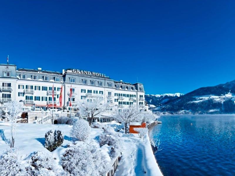 Hotel na zasněženém břehu jezera s horami v pozadí a jasnou modrou oblohou