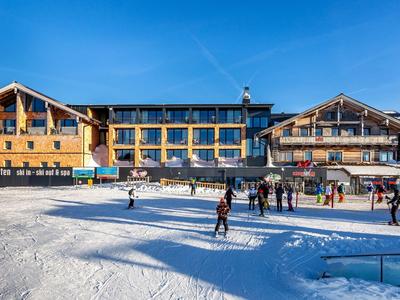 Edificio dell'hotel con facciata in legno davanti a zona sciistica innevata, persone in primo piano sotto cielo soleggiato.