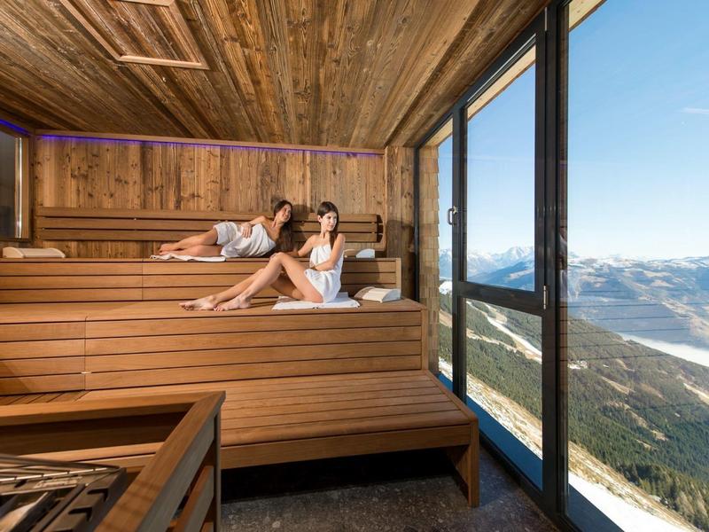 Deux femmes se détendent dans un sauna en bois avec une grande baie vitrée et vue sur la montagne.