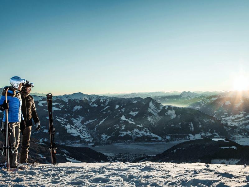 Deux randonneurs avec bâtons de trekking sur une montagne enneigée face à un paysage montagneux ensoleillé.