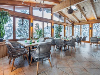 Helles Restaurant mit Holzbalken, großen Fenstern und modernen Stühlen mit Pflanzen.