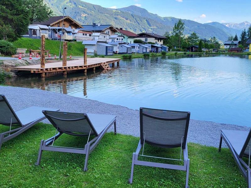 Vista de un lago tranquilo con cuatro tumbonas en el césped y un muelle al fondo.
