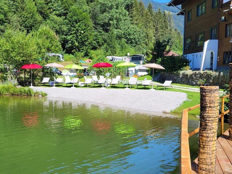 Lago con vegetación verde, terraza con sombrillas y cerca de madera junto a edificio de hotel.