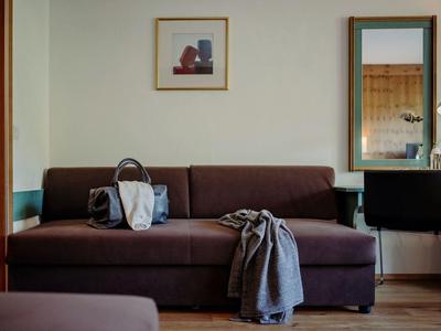 Braunes Sofa mit Tasche und Decke, Wand mit zwei gerahmten Bildern, rechts ein Schreibtisch und Stuhl.