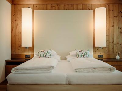 Modernes Doppelbett mit weißen Bettdecken, Holzwand und zwei beleuchteten Wandlampen.