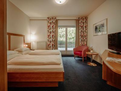 Helles Hotelzimmer mit Doppelbett, Blumen-Vorhängen, Sesseln und Flachbildfernseher auf Holzmöbeln.