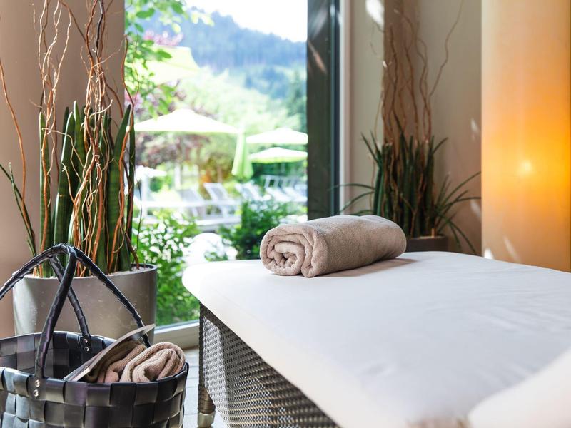 Gemütlicher Massagetisch mit beigem Handtuch, Korb und Blick auf grüne Pflanzen und Berge draußen.