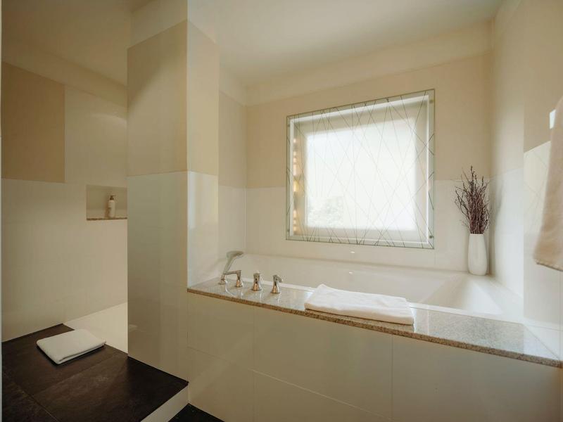 Helles Badezimmer mit Badewanne, Fenster, weißen Handtüchern und braunen Bodenfliesen.