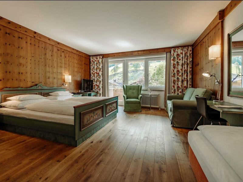 Gemütliches Schlafzimmer mit Holzdekor, Doppelbett, Sesseln, Sofa und großem Fenster mit Vorhängen.