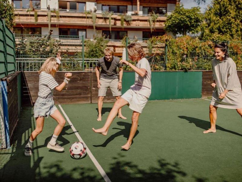 Vier Personen spielen Fußball auf einem kleinen Kunstrasenplatz neben einem Hotel