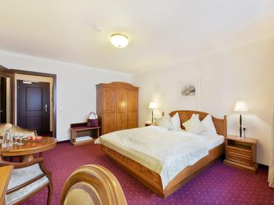 Jasno oświetlony pokój hotelowy z łóżkiem podwójnym, drewnianymi meblami i ciemnoczerwonym dywanem.