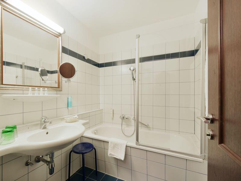 Piccolo bagno con vasca, parete doccia, lavabo e specchio con mensola.