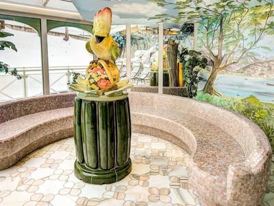 Interieur met gebogen marmeren bank, twee gele papegaaien op een groene sokkel, grote ramen en muurschildering.