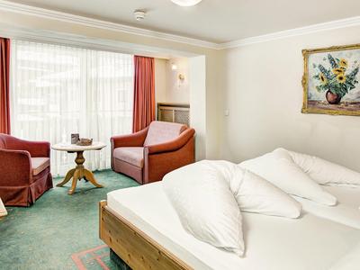 Lichte hotelkamer met tweepersoonsbed, twee fauteuils, tafel, tapijt en groot raam met gordijnen.