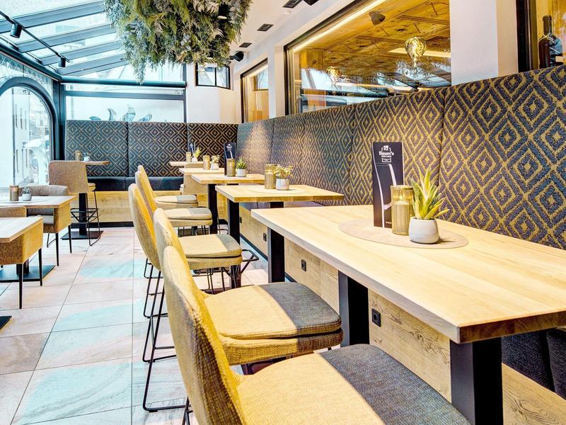 Modern café met houten stoelen, beklede banken en planten aan het plafond.