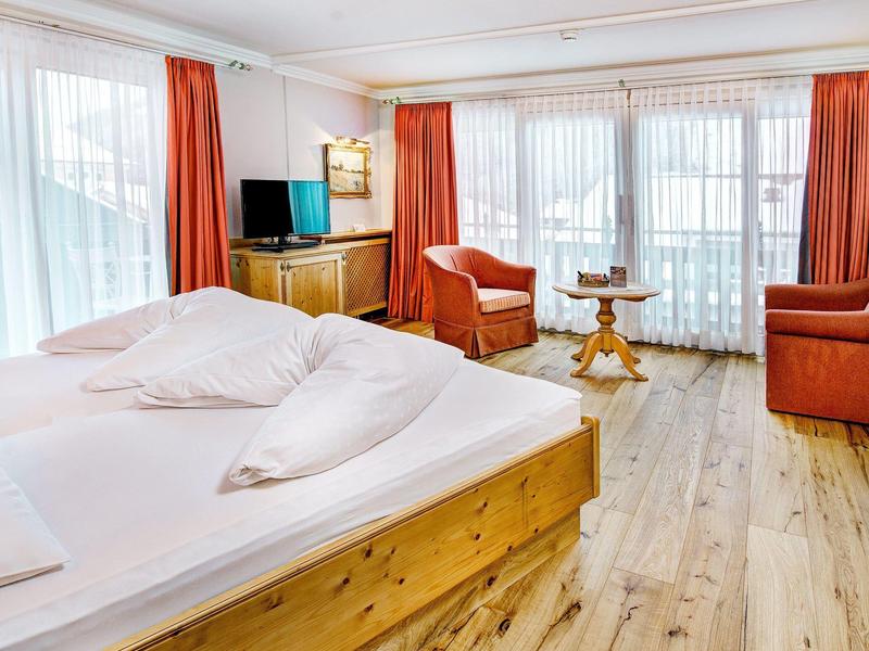 Gezellige hotelkamer met houten meubels, twee fauteuils en grote ramen met rode gordijnen.