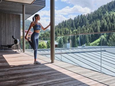 Frau in Sportbekleidung steht auf Holzterrasse mit Blick auf Wald und Berge.