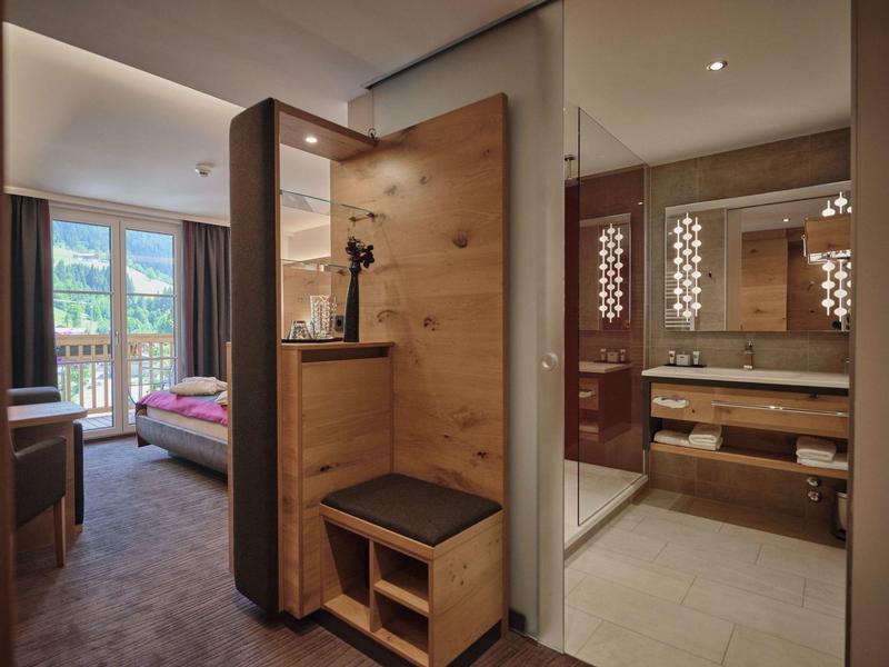 Moderne hotelkamer met houten meubels, balkonuitzicht en aangrenzende badkamer met grote spiegels.