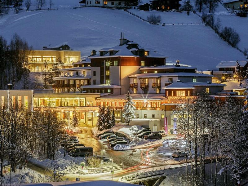Verlicht hotel in besneeuwd winterlandschap 's nachts met omliggende bomen.