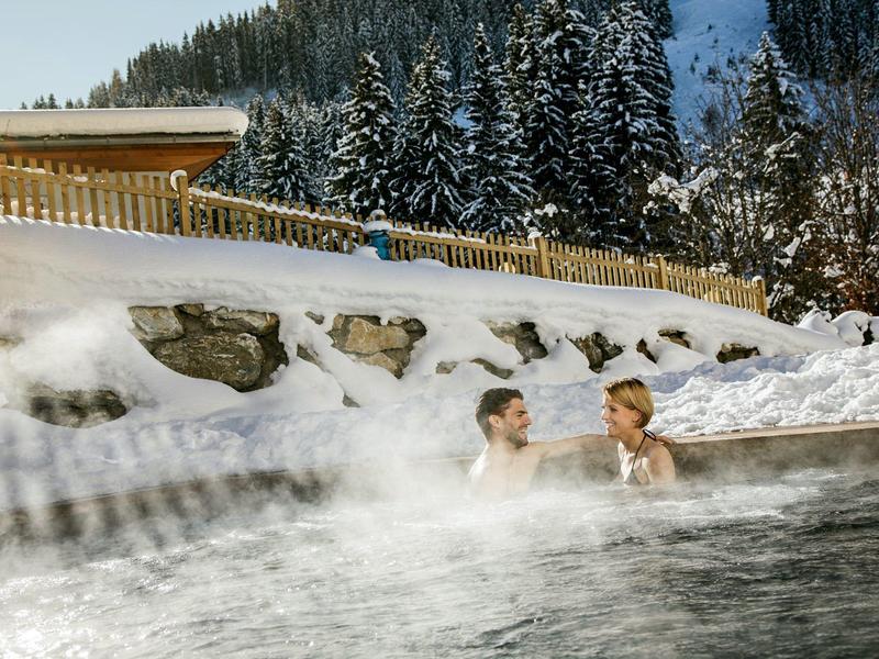 Twee mensen ontspannen in een buitenjacuzzi omringd door sneeuw en dennenbomen.