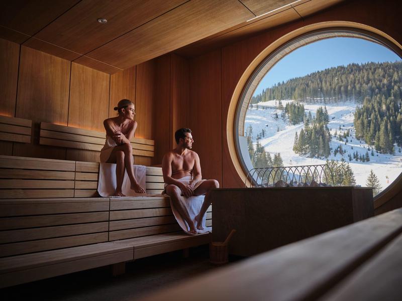 Twee mensen ontspannen in een sauna met een groot rond raam met uitzicht op besneeuwde bergen.