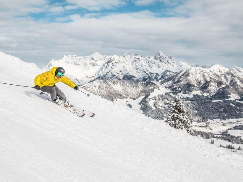 Esquiador con chaqueta amarilla baja por una pendiente nevada con panorama montañoso.