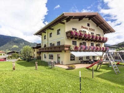 Gelbes Haus mit Blumen an Balkonen, großem grünen Rasen und Spielplatz, Berge im Hintergrund.