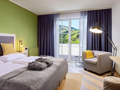 Nowoczesny pokój hotelowy z podwójnym łóżkiem, zieloną ścianą i balkonem z widokiem na góry.