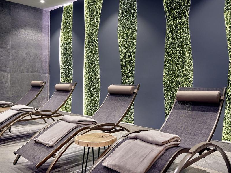 Nowoczesne leżaki z poduszkami i kocami przed dekoracyjną ścianą z zielonymi pionowymi pasami roślin.