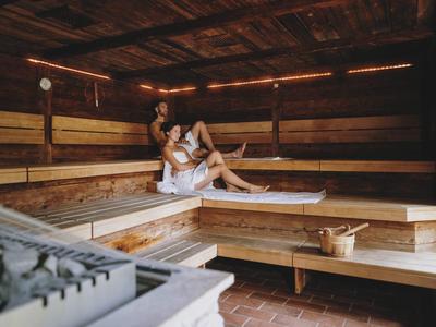 Dvě osoby odpočívají na lavicích v sauně z dřevěných trámů s vědrem na saunu.