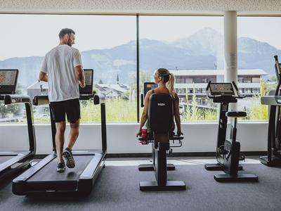 A man runs on a treadmill and a woman exercises on a bike with mountain views in the background.