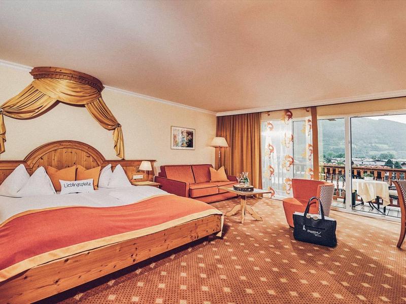 Prostorný hotelový pokoj s velkou postelí, kobercem, sezením a balkonem s výhledem na hory.