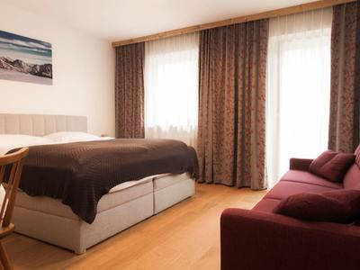 Chambre lumineuse avec lit double, sol en bois, et canapé rouge près de la fenêtre avec rideaux.