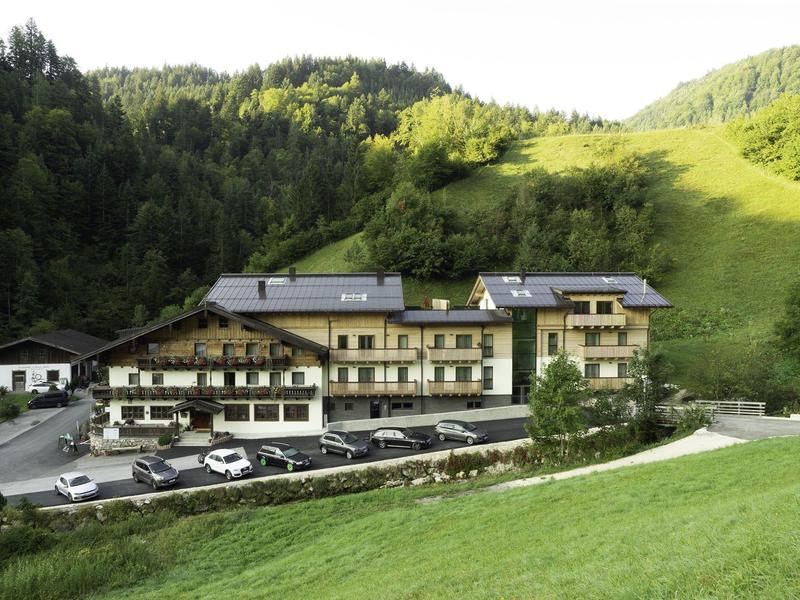 Large hotel with parking lot in front of forested hill and green meadow.