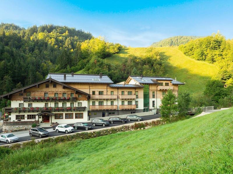 Large hotel with multiple parking spaces in front of green hills and forest in the background.