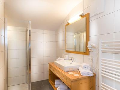 Moderní koupelna s bílým obloženým sprchovým koutem, dřevěnou umyvadlovou skříňkou a osvětleným zrcadlem.