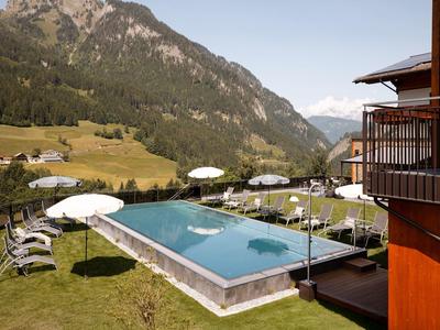 Außenpool mit Liegen und Sonnenschirmen auf einer Wiese vor Bergkulisse bei klarem Himmel.