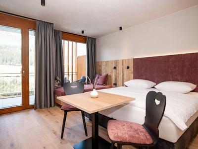 Moderně zařízený hotelový pokoj s manželskou postelí, dřevěným stolem a balkonem s výhledem na hory.