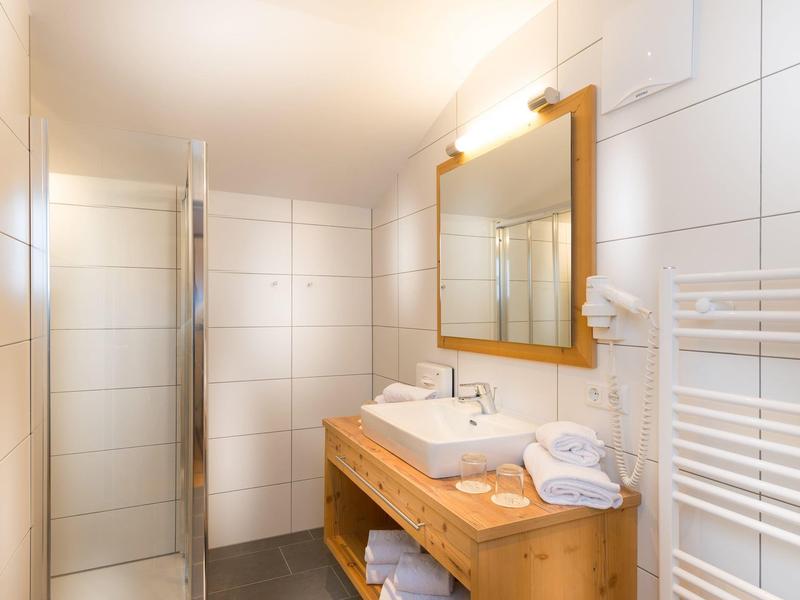 Moderní koupelna s bílým obloženým sprchovým koutem, dřevěnou umyvadlovou skříňkou a osvětleným zrcadlem.