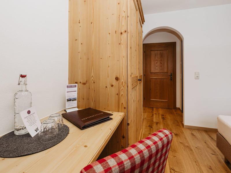 Dřevěný stůl se skleněnou lahví s vodou, červenou kostkovanou židlí a dřevěnou skříní v světlé místnosti.