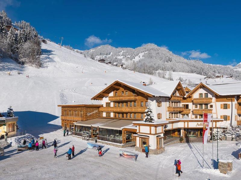 Station d'hiver avec bâtiments en bois et skieurs devant des montagnes enneigées sous un ciel bleu.
