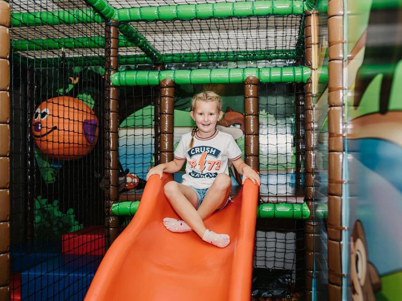Enfant assis sur un toboggan orange dans une aire de jeux intérieure avec des structures d'escalade.