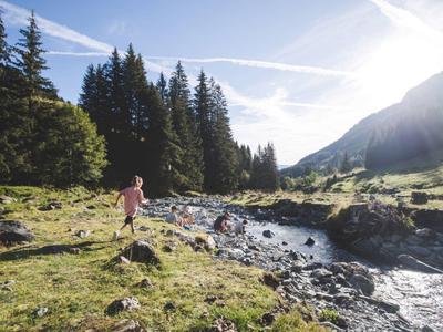 Un paysage avec une montagne, une forêt et un petit ruisseau où deux enfants jouent près de la prairie.