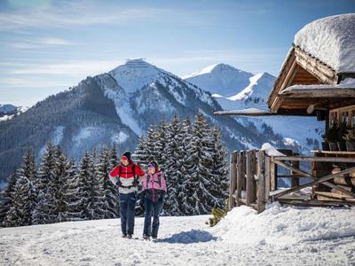 Dvě osoby v zimním oblečení kráčejí ve sněhu vedle dřevěné chaty v horách.