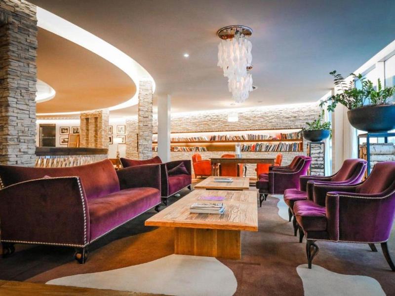 Moderní hotelový salonek s fialovými křesly, pohovkou, dřevěným stolem a elegantním lustrem.