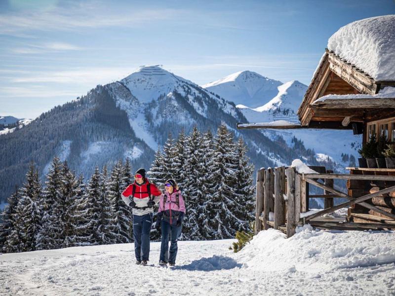 Dvě osoby v zimním oblečení kráčejí ve sněhu vedle dřevěné chaty v horách.