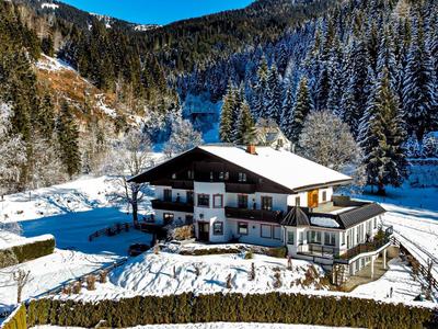Vecchio chalet in una valle innevata circondata da foresta e montagne sullo sfondo.