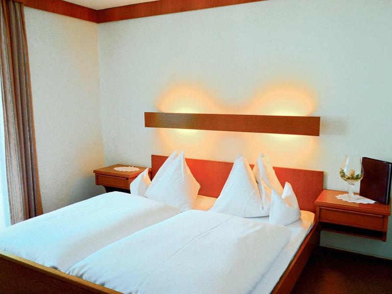 Světle zařízený hotelový pokoj s manželskou postelí, bílými polštáři a nočními stolky.